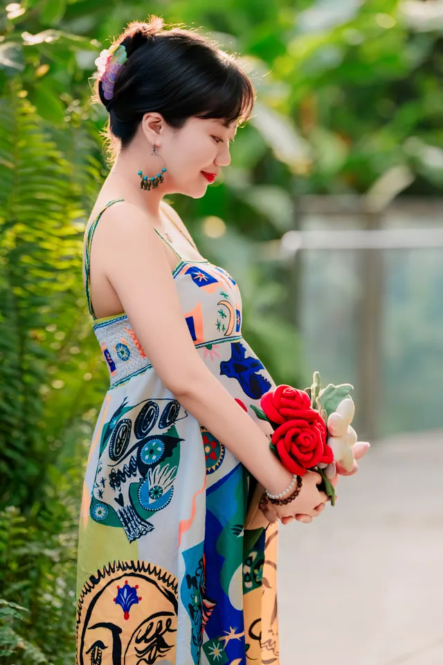 穿花纹长裙的准妈妈侧身捧着红玫瑰，背景是庭院绿植